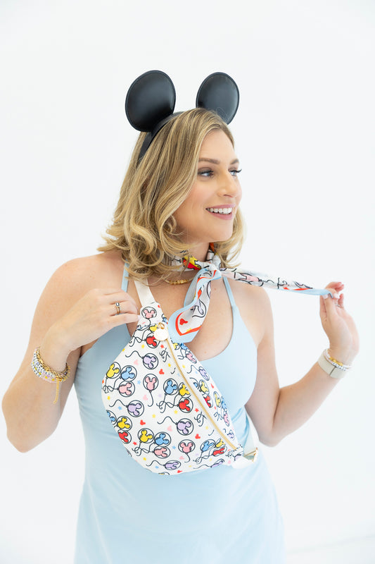 A woman wearing black Mouse ears and a light blue dress models a white fanny pack with a colorful cartoon print. She is holding the tie of the bag, which features a gold zipper, against a plain white background.