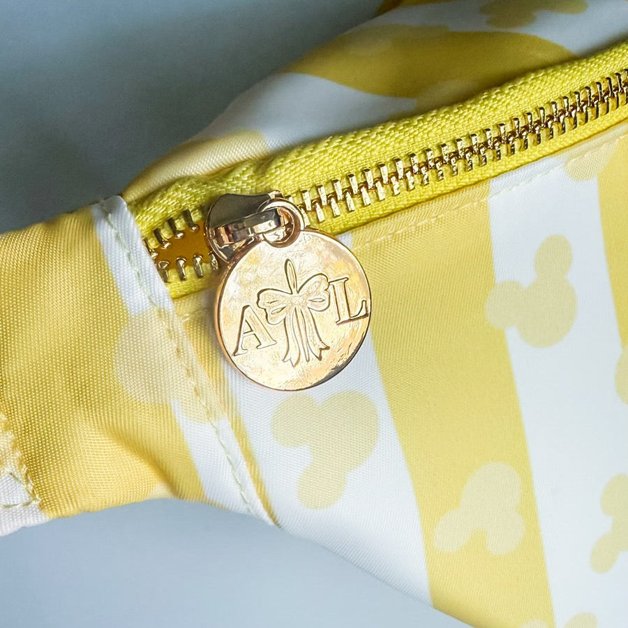 Close-up of a yellow and white patterned bag with a gold zipper and decorative tag on a light blue background.