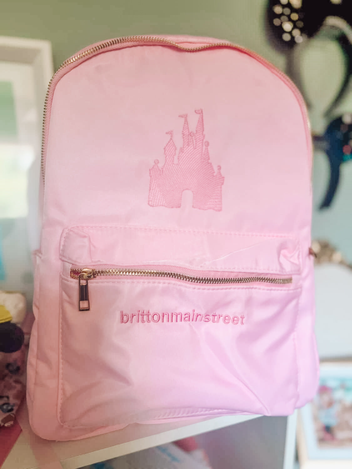 Pink backpack with castle design and 'brittonmainstreet' branding on a blurred background