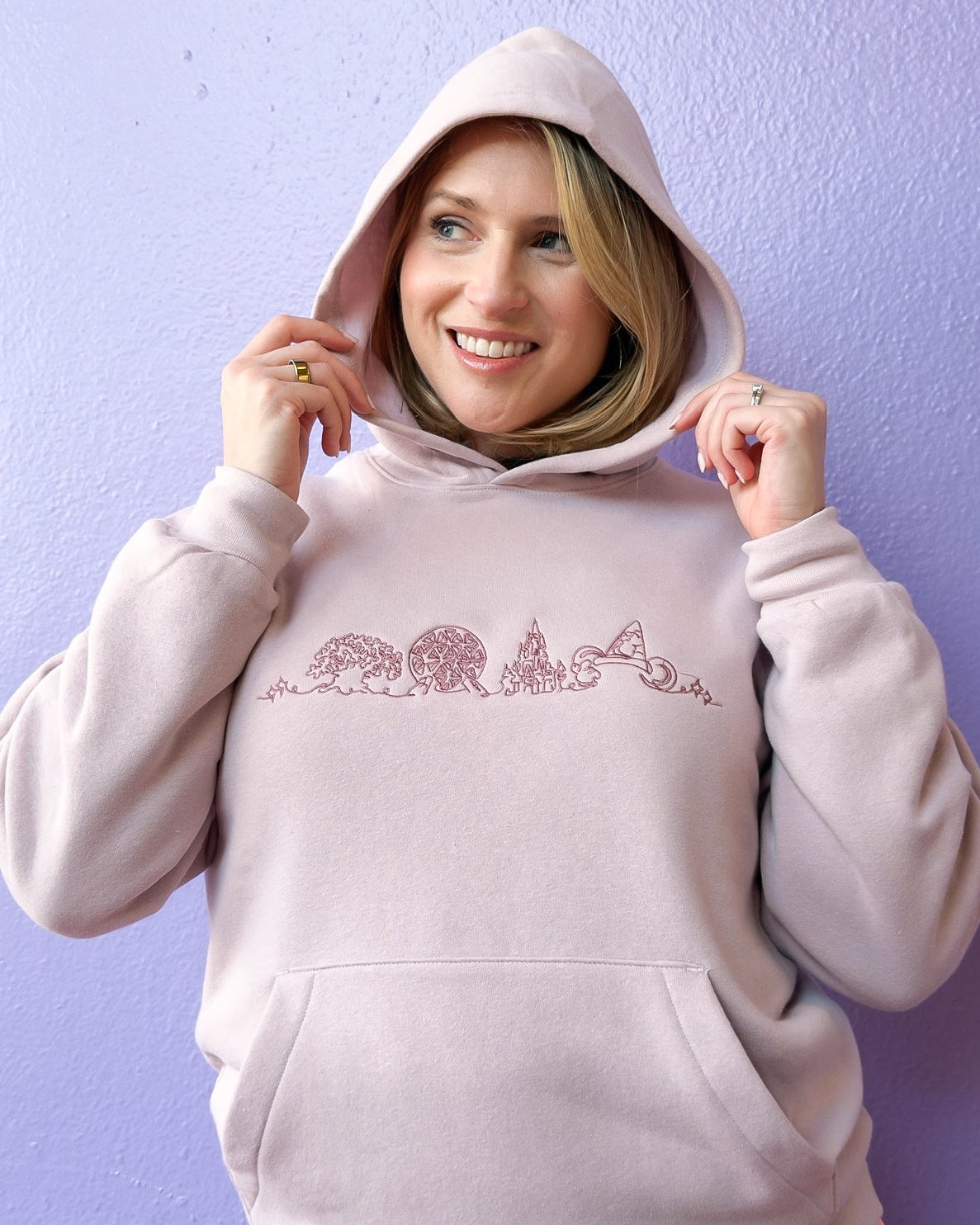 Woman wearing a light gray hoodie with a design and a pink pleated skirt against a purple background