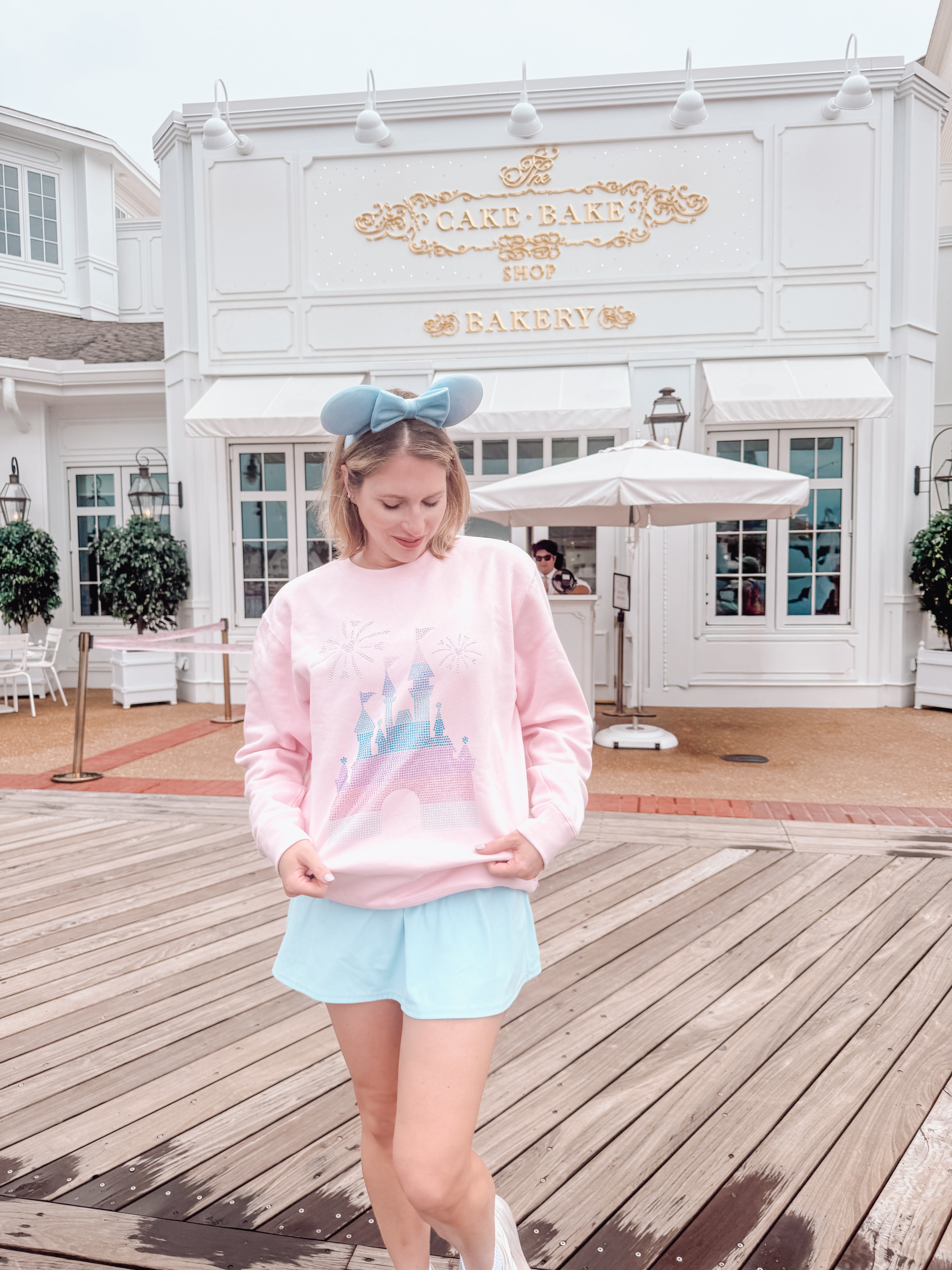Woman wearing a pink sweatshirt with a blue bow in front of a bakery.