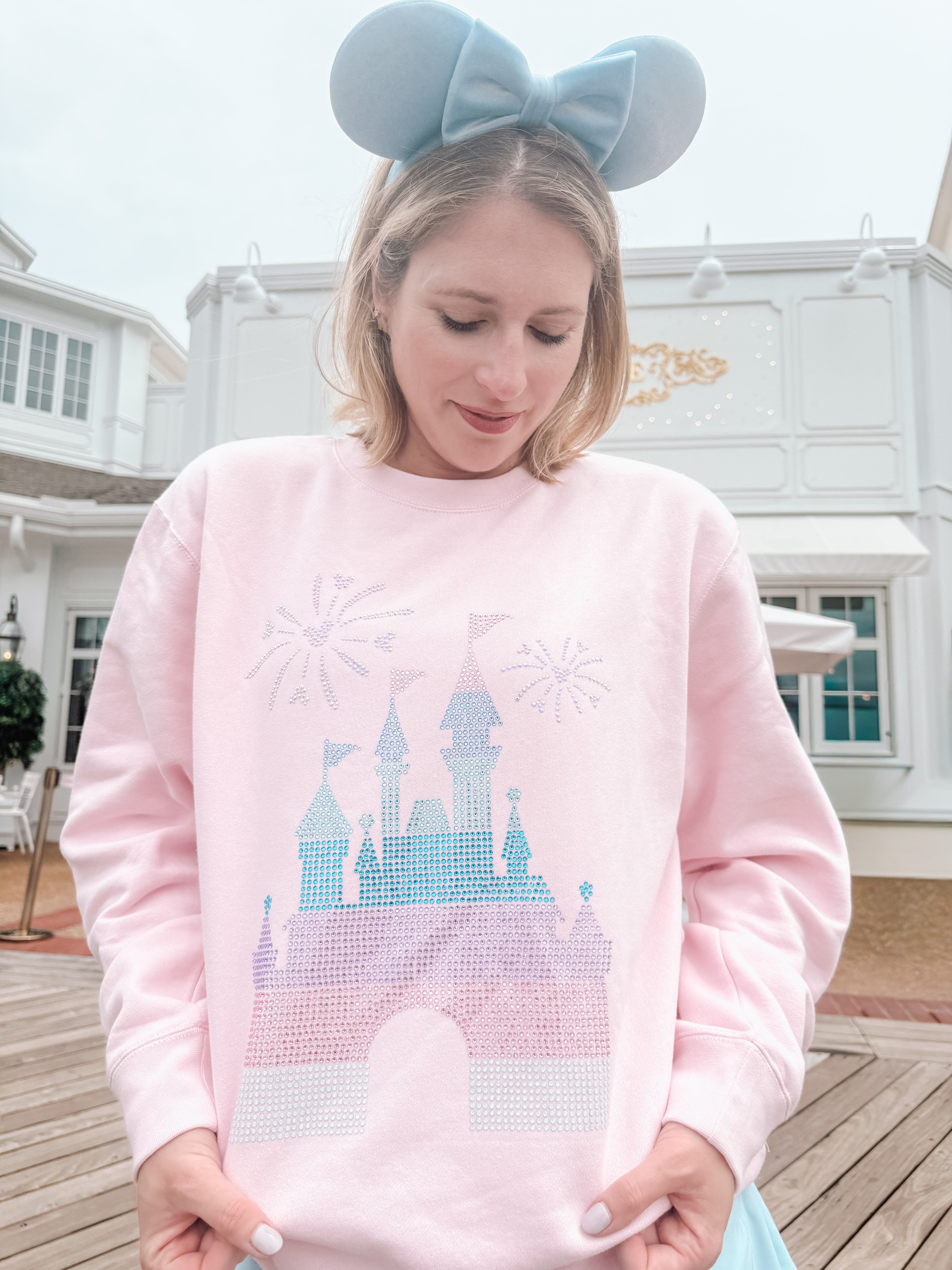 Person wearing a pink sweatshirt with a castle design and blue mouse ears in front of a building.