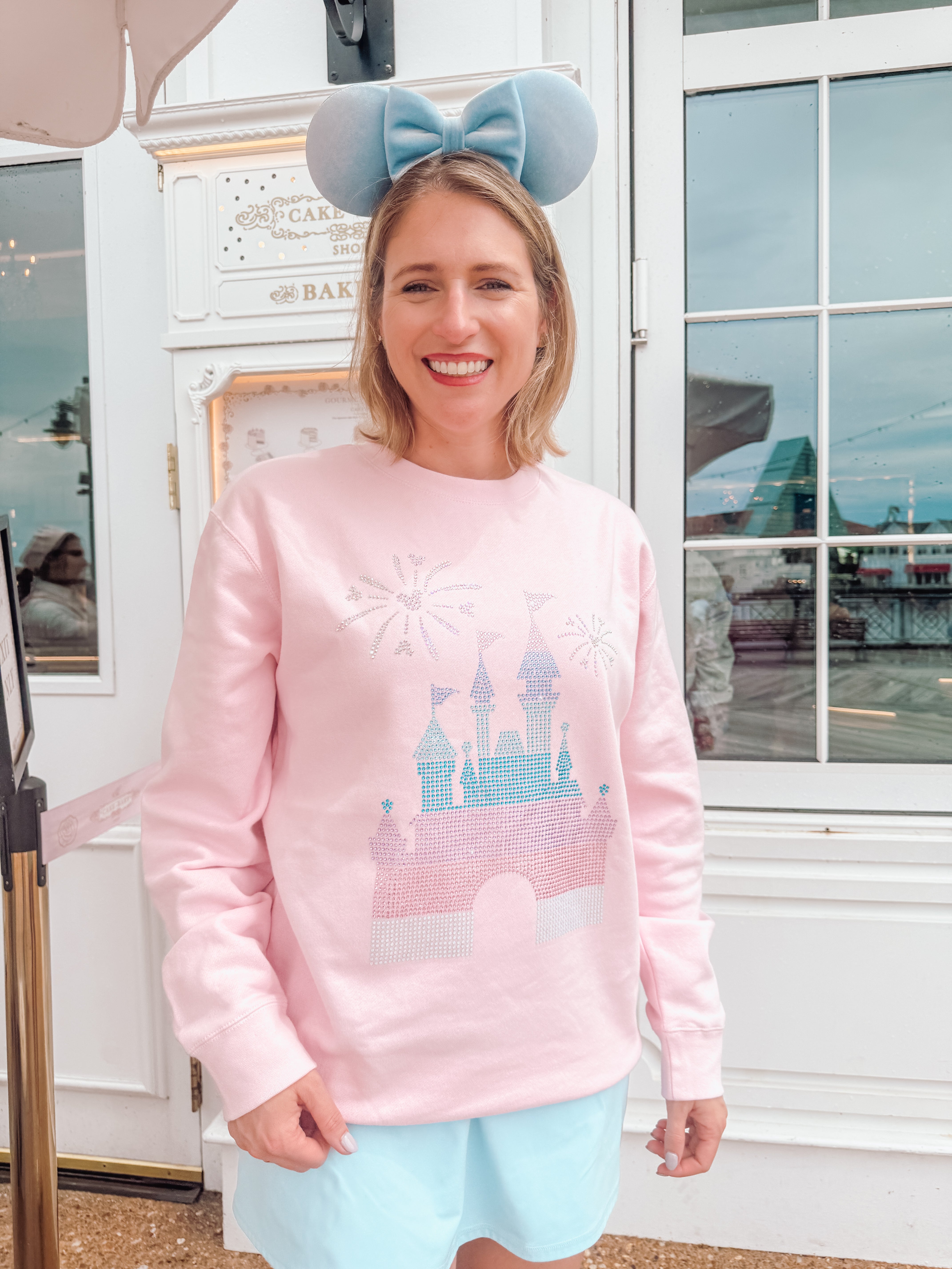 Person wearing a pink sweatshirt with a castle design and light blue shorts, standing indoors.