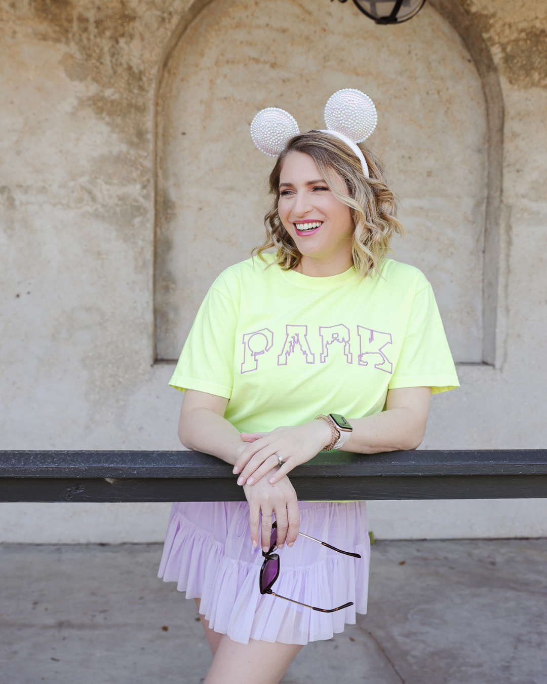 Woman wearing a bright yellow 'PArk' t-shirt and light purple skirt with white mouse ears, leaning on a railing.
