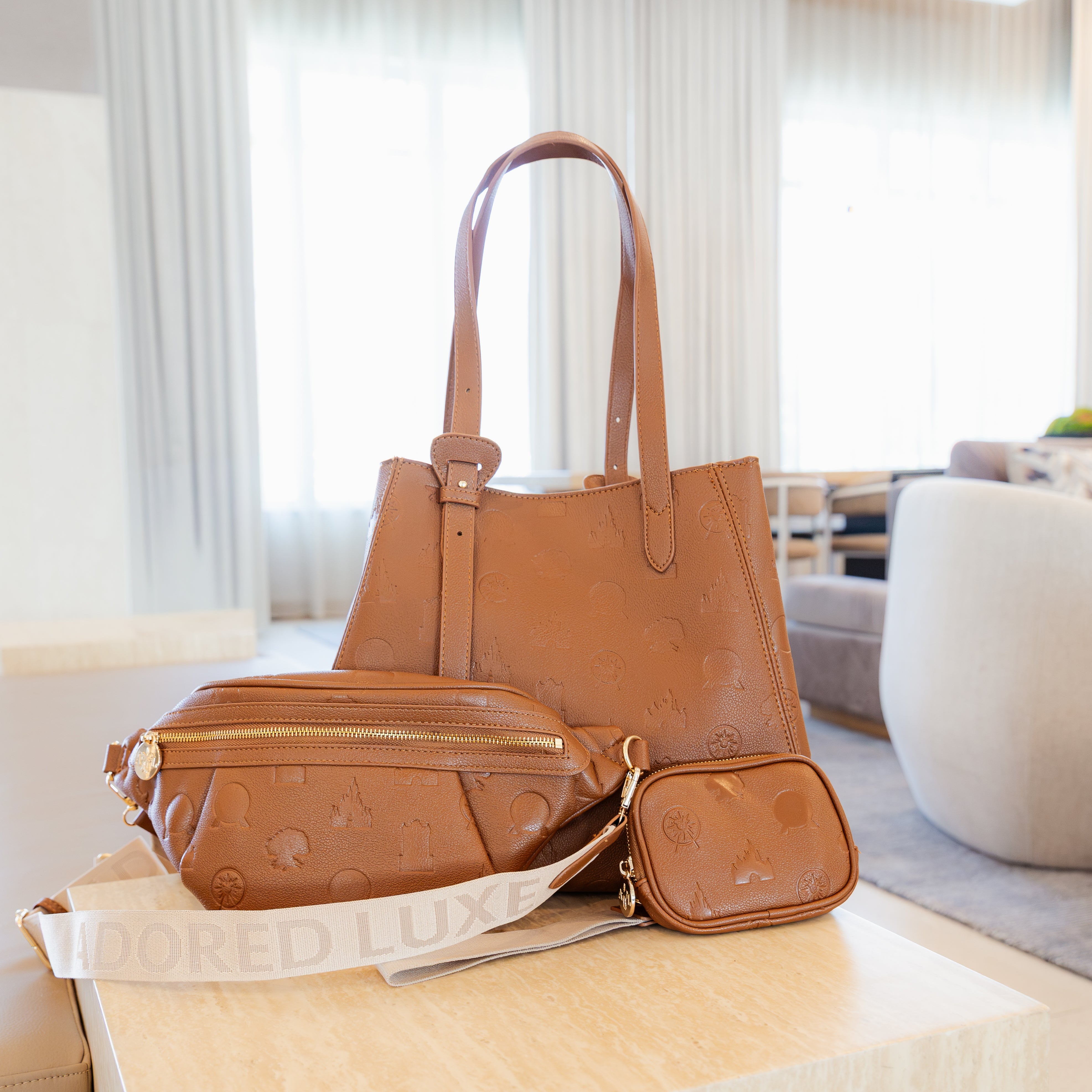 A tan leather handbag set displayed on a light wood table in a bright, modern living room. The set includes a large tote bag, a matching crossbody bag with a white embossed strap, and two small zip-around pouches.