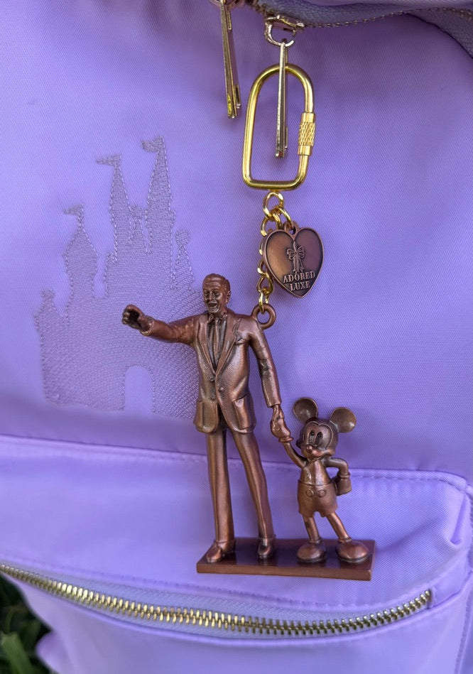 Bronze keychain of a man and a mouse holding hands on a purple surface.