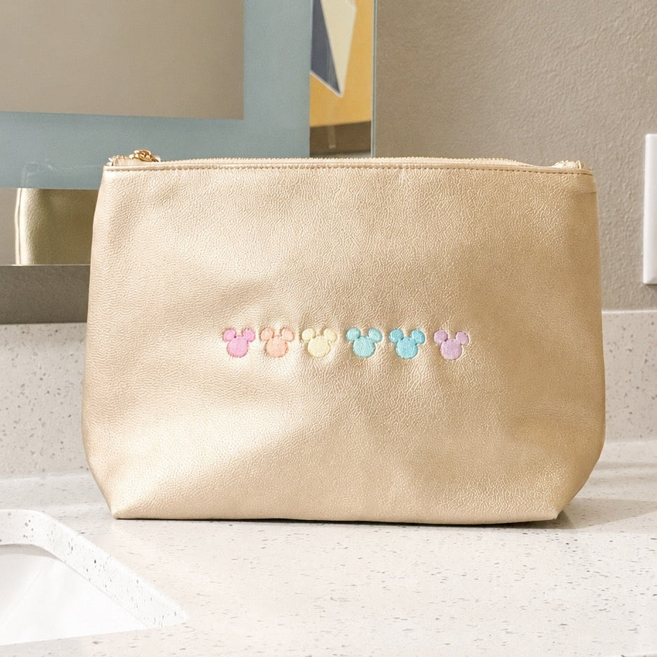 A gold faux-leather cosmetic pouch with a small embroidered row of colorful Mickey Mouse head silhouettes on the front, sitting on a bathroom countertop.