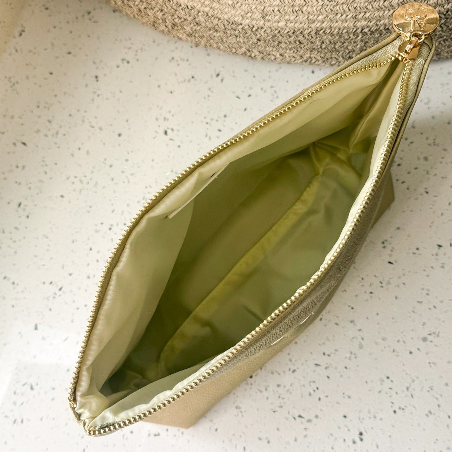 A metallic gold zippered pouch with an ivory interior, shown open on a white speckled countertop next to a woven basket.