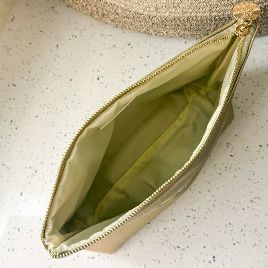 A metallic gold zippered pouch with an ivory interior, shown open on a white speckled countertop next to a woven basket.