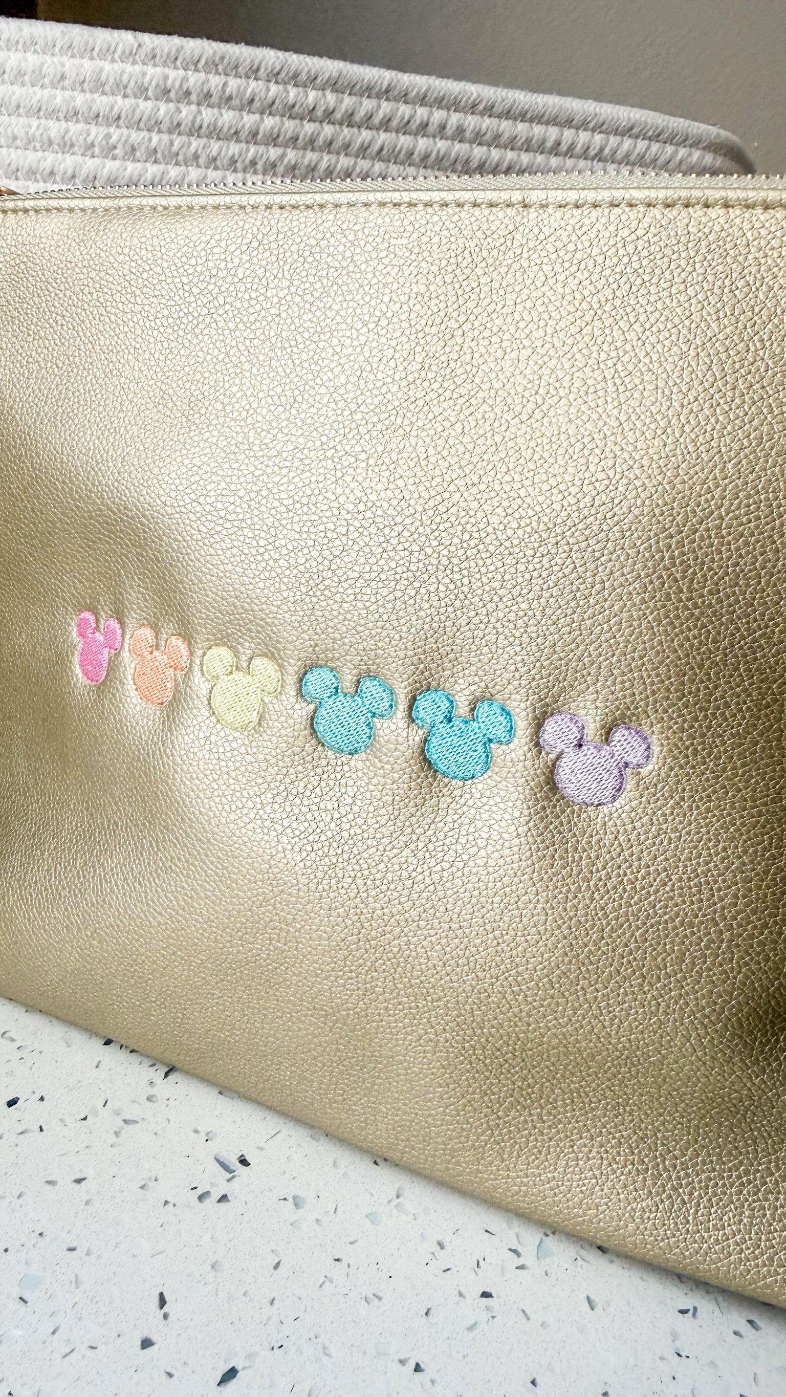 A close-up of a metallic gold textured pouch with five small pastel embroidered Mickey Mouse head silhouettes across the front, resting on a speckled white surface.
