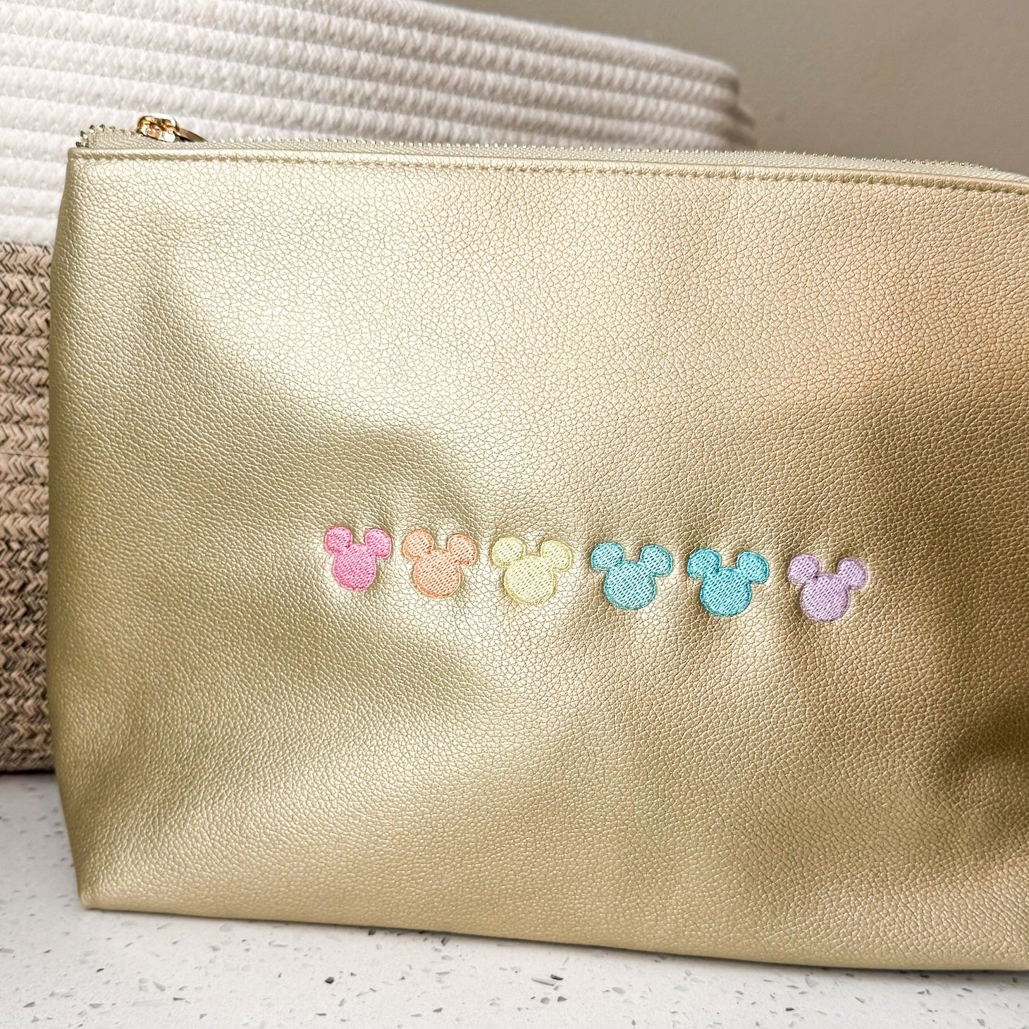 A metallic gold faux-leather pouch with a row of small pastel embroidered Mickey Mouse head silhouettes across the front, resting on a speckled white surface.