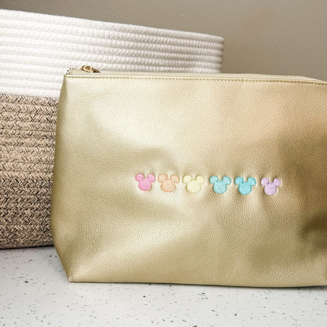A metallic gold cosmetic pouch with a row of small pastel Mickey Mouse head charms, resting on a speckled white countertop next to a woven basket.
