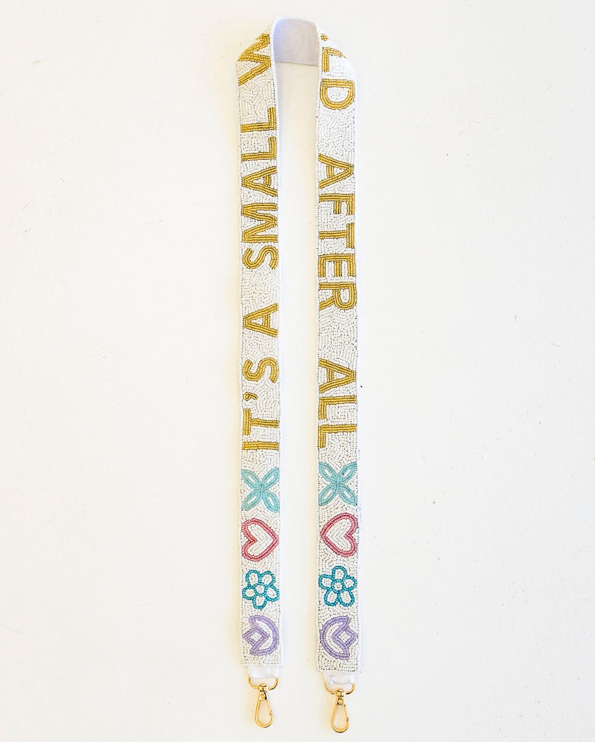 A white beaded purse strap with gold text reading 'IT'S A SMALL WORLD AFTER ALL' and decorated with pastel pink, blue, and purple hearts, flowers, and bows, shown on a plain white background.